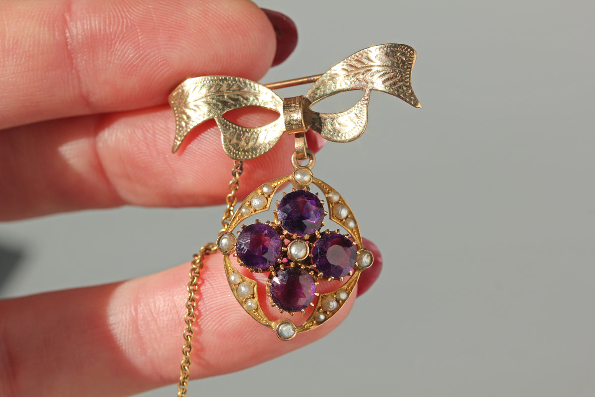 A 9ct gold brooch shaped like a bow with a dangling floral design set with purple amethyst gemstones and small white pearls, held by a hand against a neutral gray background.