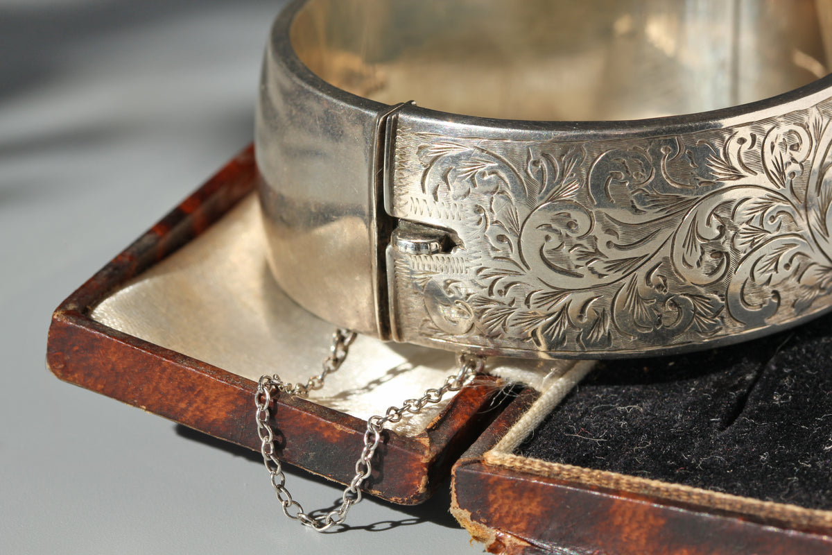 An ornate engraved silver cuff bracelet with a chain, displayed on top of an open vintage brown leather case.