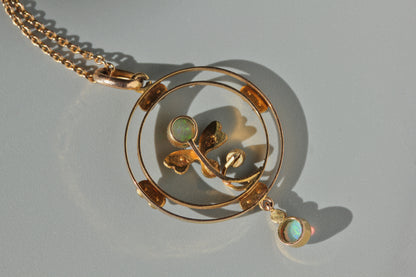 Hallmark close up of 9ct gold pendant necklace featuring a circular double-ring design with a stylized leaf motif and two small iridescent opal-like gemstone accents, displayed on a neutral gray surface.
