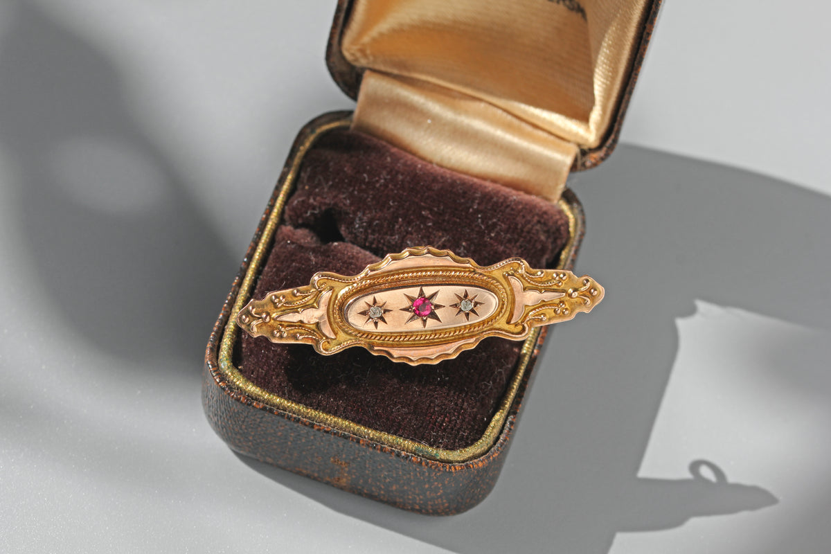 A victorian 9ct gold brooch with star and starburst enamel details, displayed inside an open vintage jewelry box with a gold interior and dark brown velvet lining.