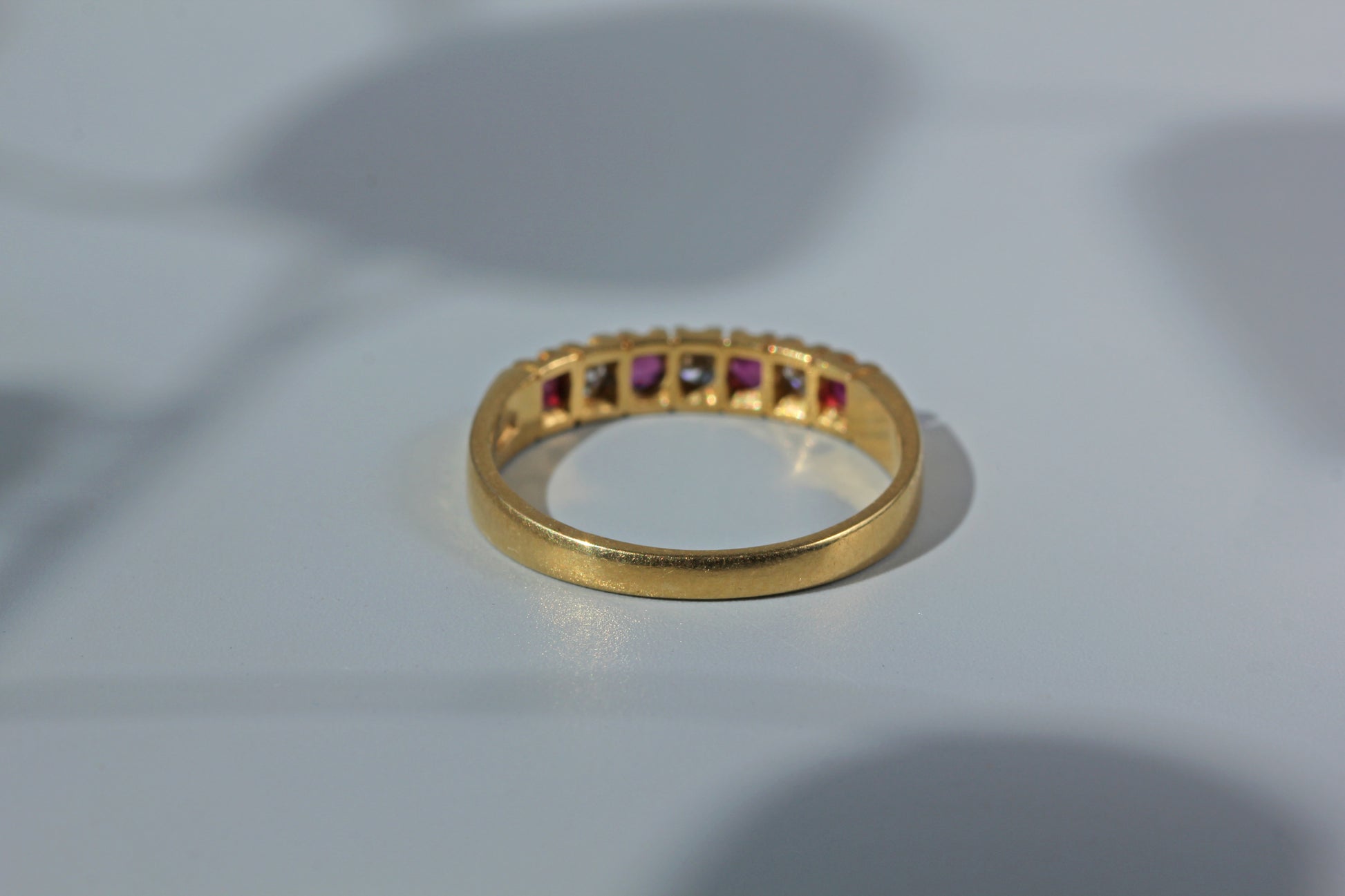 A gold band ring featuring a row of small square-cut gemstones in alternating pink, clear, and dark tones, resting on a light gray surface.