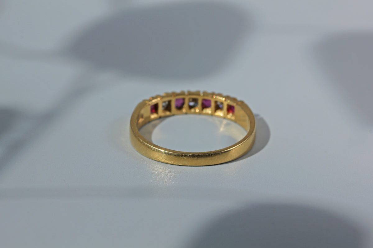 A gold band ring featuring a row of small square-cut gemstones in alternating pink, clear, and dark tones, resting on a light gray surface.