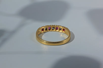 A gold band ring featuring a row of small square-cut gemstones in alternating pink, clear, and dark tones, resting on a light gray surface.