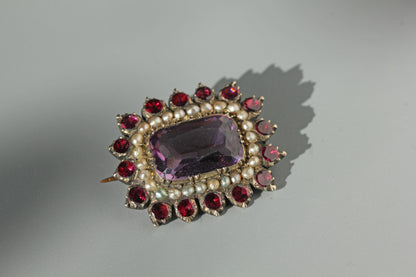 Antique 9ct gold Georgian brooch with cushion cut amethyst, seed pearls, flat cut garnets and a spiky floral silhouette displayed on a neutral background 