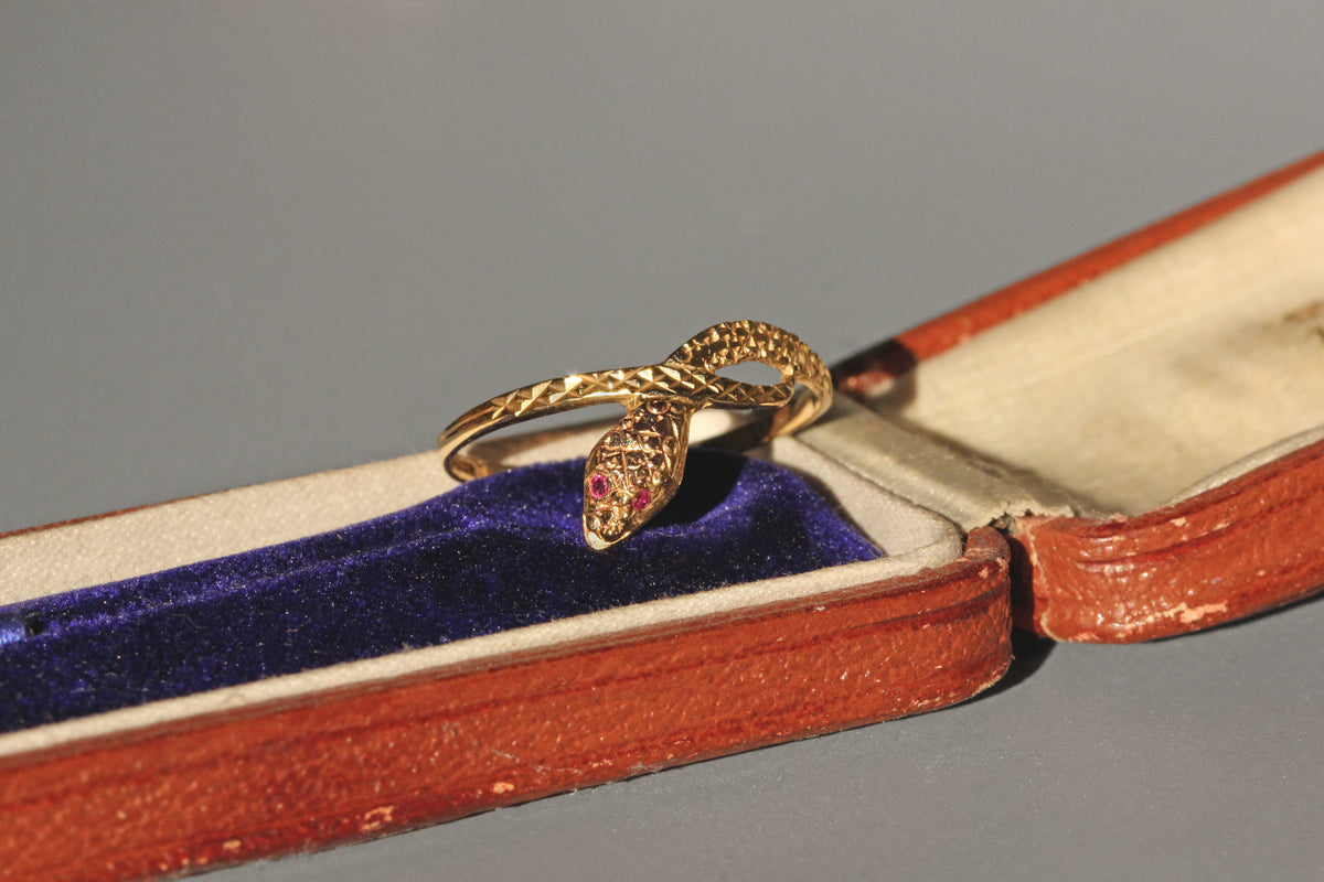 Gold snake-shaped ring with ruby eyes in an open jewelry box on a gray background