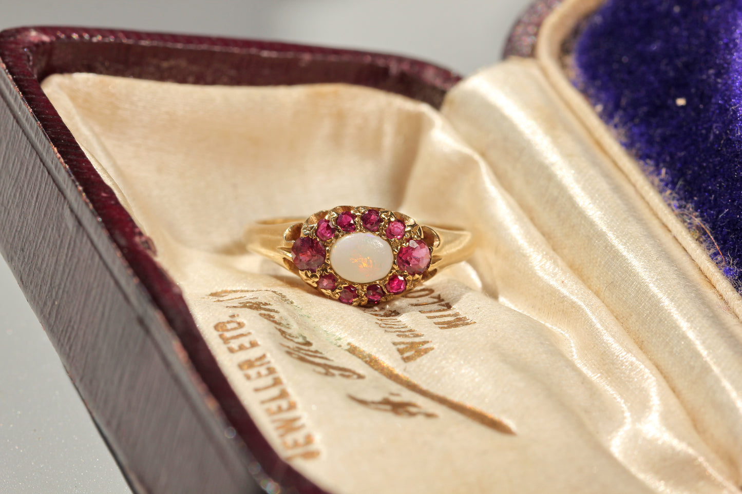 A vintage yellow gold ring with a central white opal surrounded by pink gemstones, displayed inside an antique velvet jewelry box.