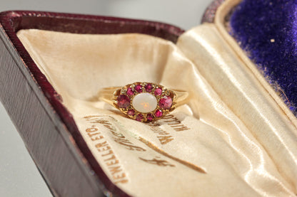 A vintage yellow gold ring with a central white opal surrounded by pink gemstones, displayed inside an antique velvet jewelry box.