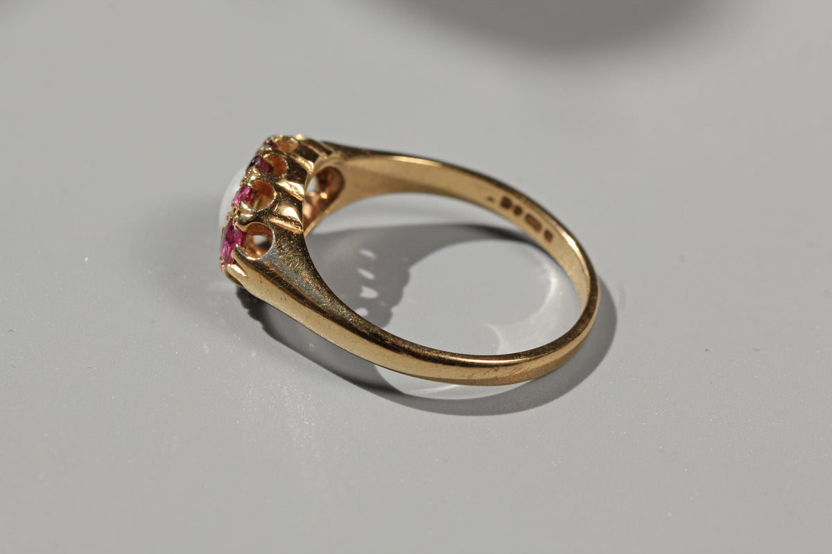 A yellow gold ring with a decorative crown-like top set with small pink gemstones, photographed on a light gray surface.