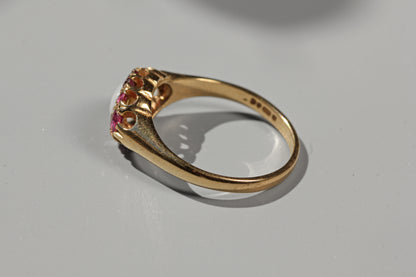 A yellow gold ring with a decorative crown-like top set with small pink gemstones, photographed on a light gray surface.
