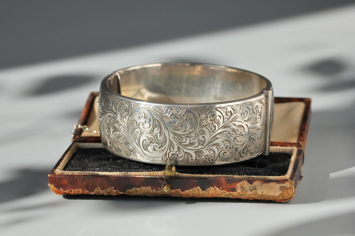 An ornate vintage sterling silver cuff bracelet with intricate floral engraving, displayed on top of an antique brown leather jewelry box.