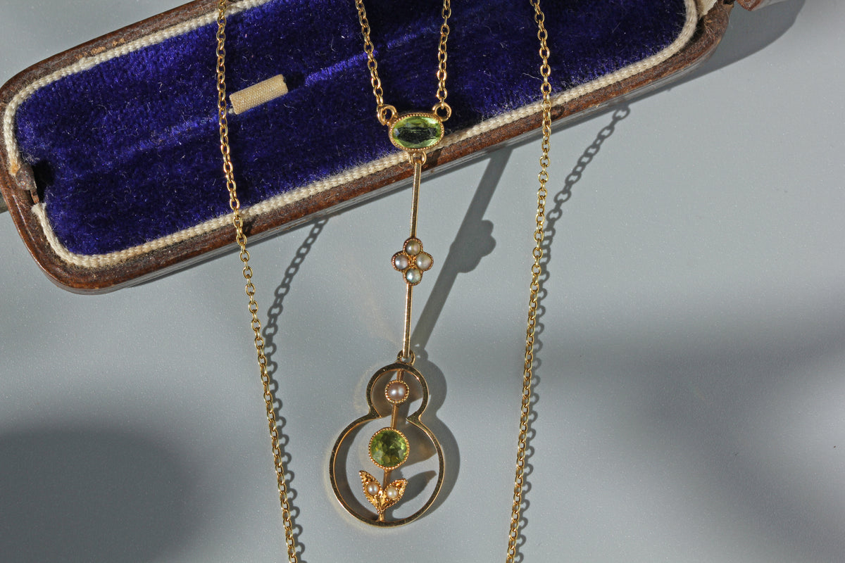 A 15ct gold antique pendant necklace with a peridot gemstone and pearl floral details, displayed in front of a blue velvet jewelry box on a light gray background.