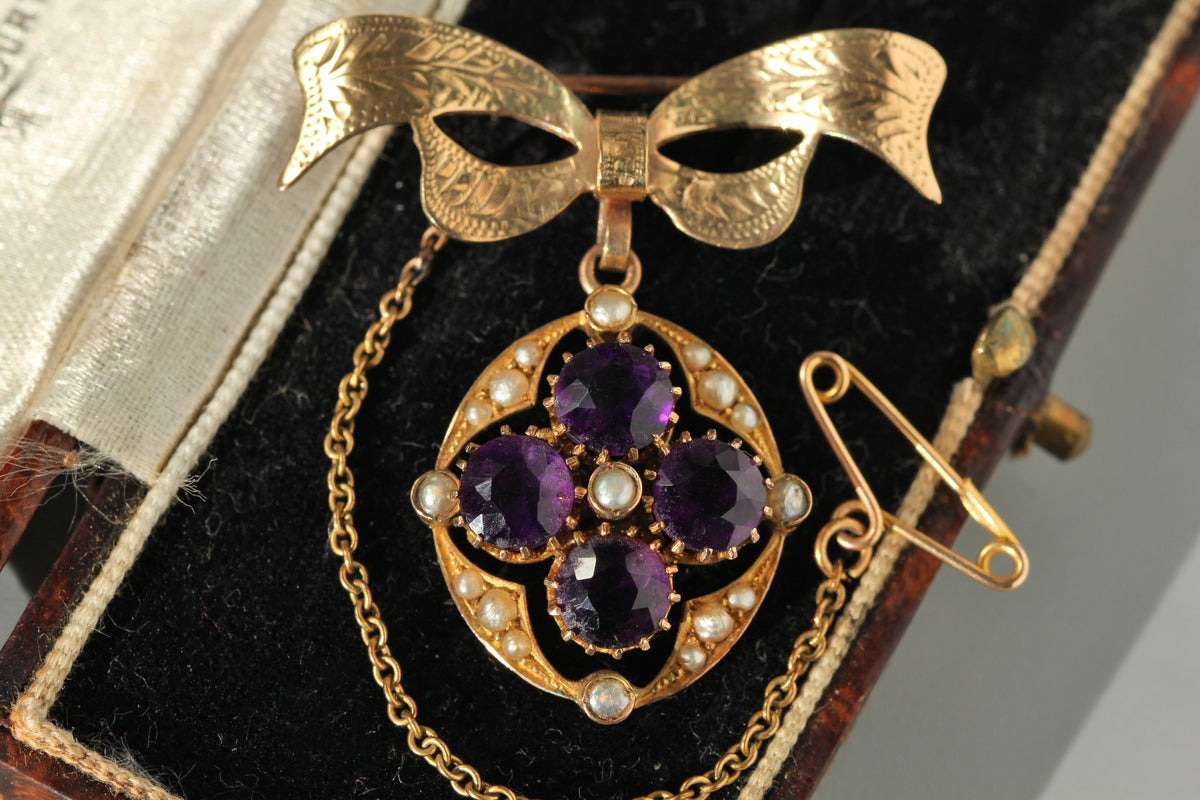 A vintage 9ct gold pendant brooch featuring four round purple amethyst gemstones in a floral arrangement, surrounded by small white pearls and a decorative bow-shaped top, displayed on a black velvet jewelry box.
