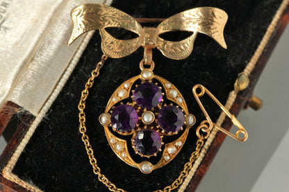 A vintage 9ct gold pendant brooch featuring four round purple amethyst gemstones in a floral arrangement, surrounded by small white pearls and a decorative bow-shaped top, displayed on a black velvet jewelry box.