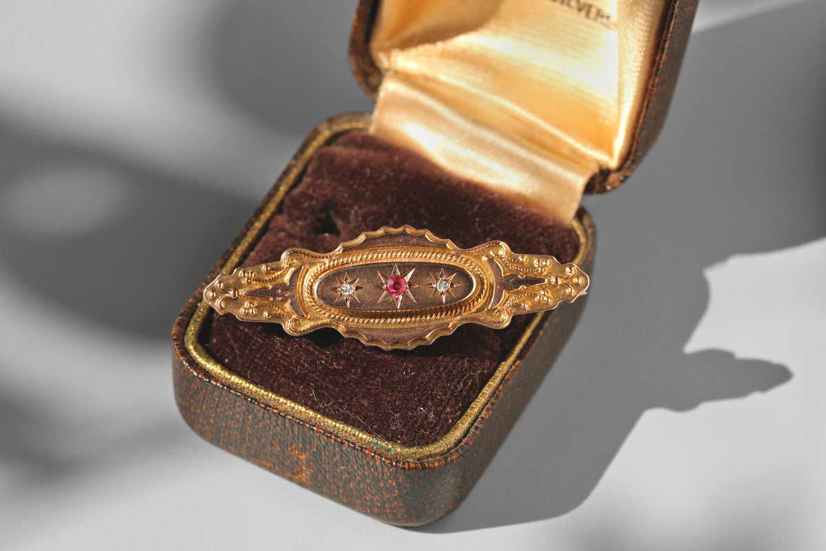An ornate 9ct gold Etruscan victorian brooch with a central pink spinel gemstone and small diamonds, displayed inside an open vintage brown velvet jewelry box with a gold satin lining on a gray background.