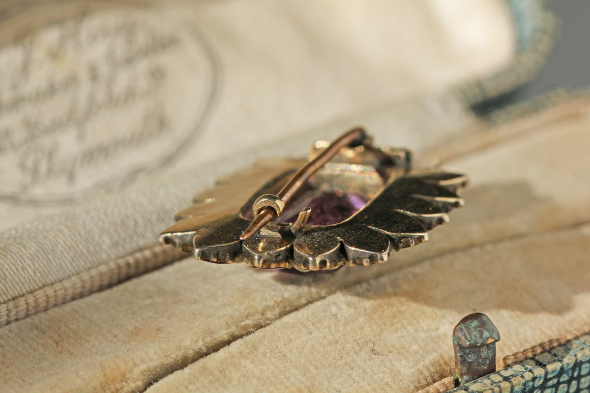 A close-up of a Georgian 9ct gold brooch with a scalloped leaf or wing-like outline and a purple faceted stone, resting on a beige fabric-lined box.