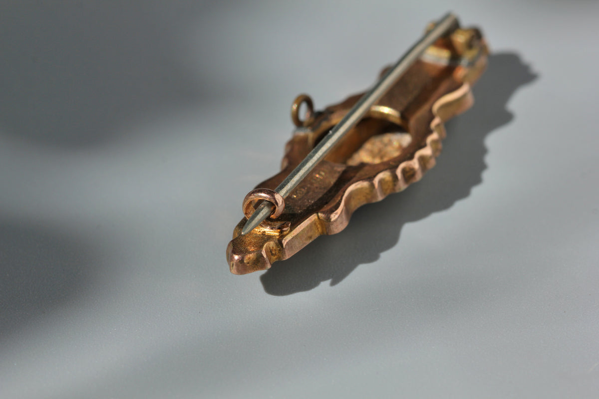 An ornate vintage 9ct brooch with a decorative wavy border and a pin bar, photographed on a neutral gray surface with a soft shadow.