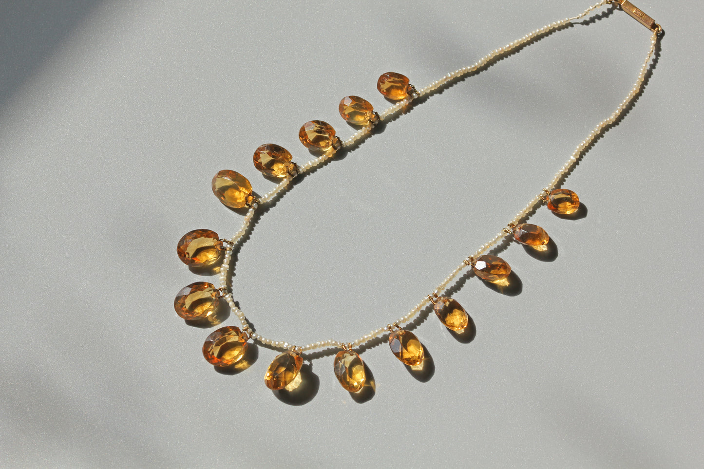 A beaded necklace featuring multiple faceted amber-yellow teardrop gemstones set in a light beige or off-white beaded chain, photographed on a neutral light gray background with soft shadows.