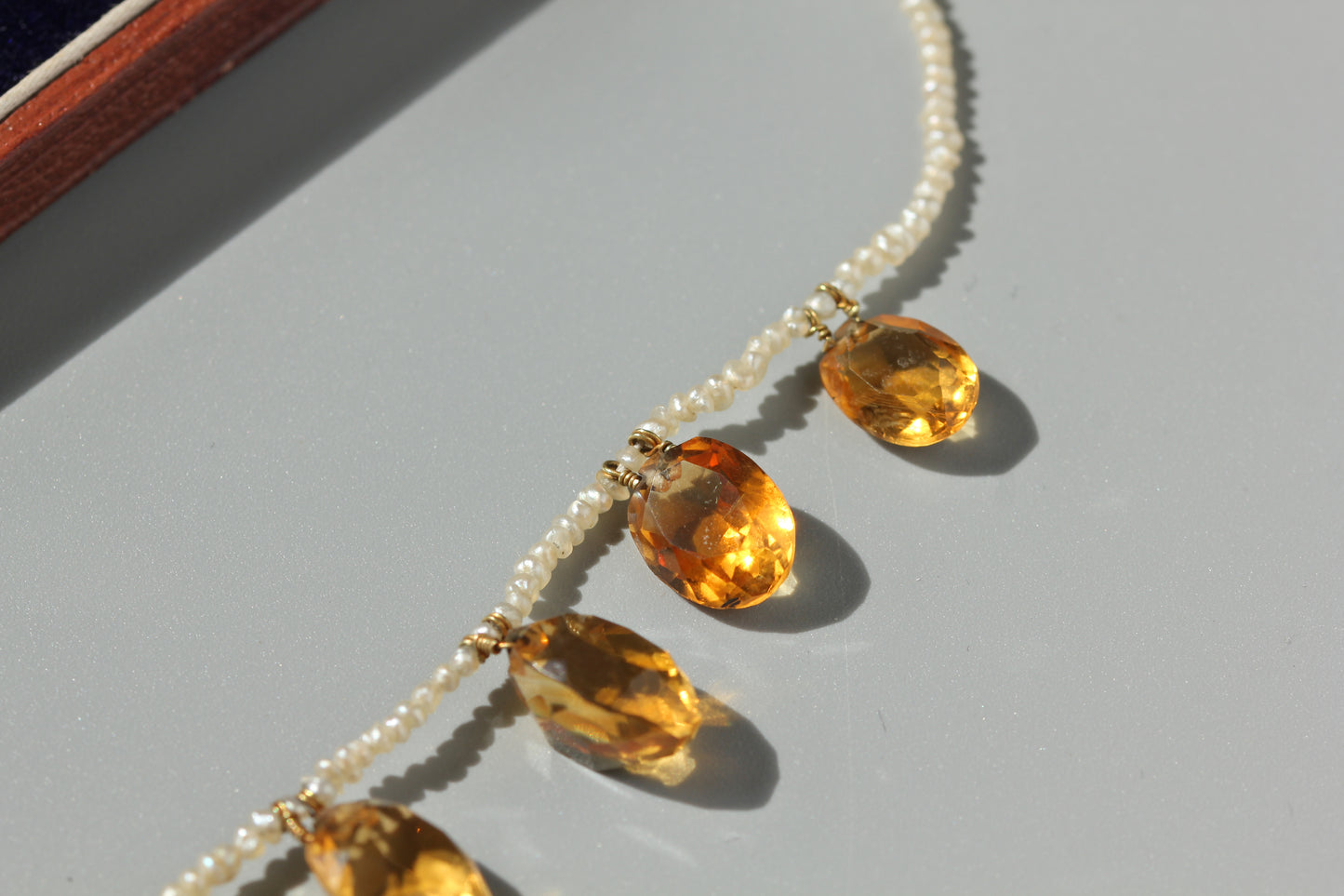 A close-up of a beaded necklace featuring small white pearls and large faceted amber-orange oval gemstones, photographed on a light gray surface with strong sunlight and shadows.