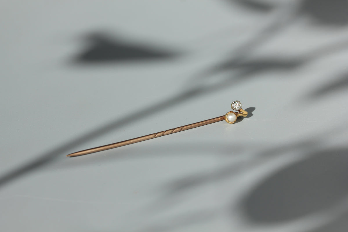 An 18ct gold art deco pin with a twisted stem and a decorative top featuring an old cut diamond and a white pearl, resting on a light gray surface with soft leaf shadows.