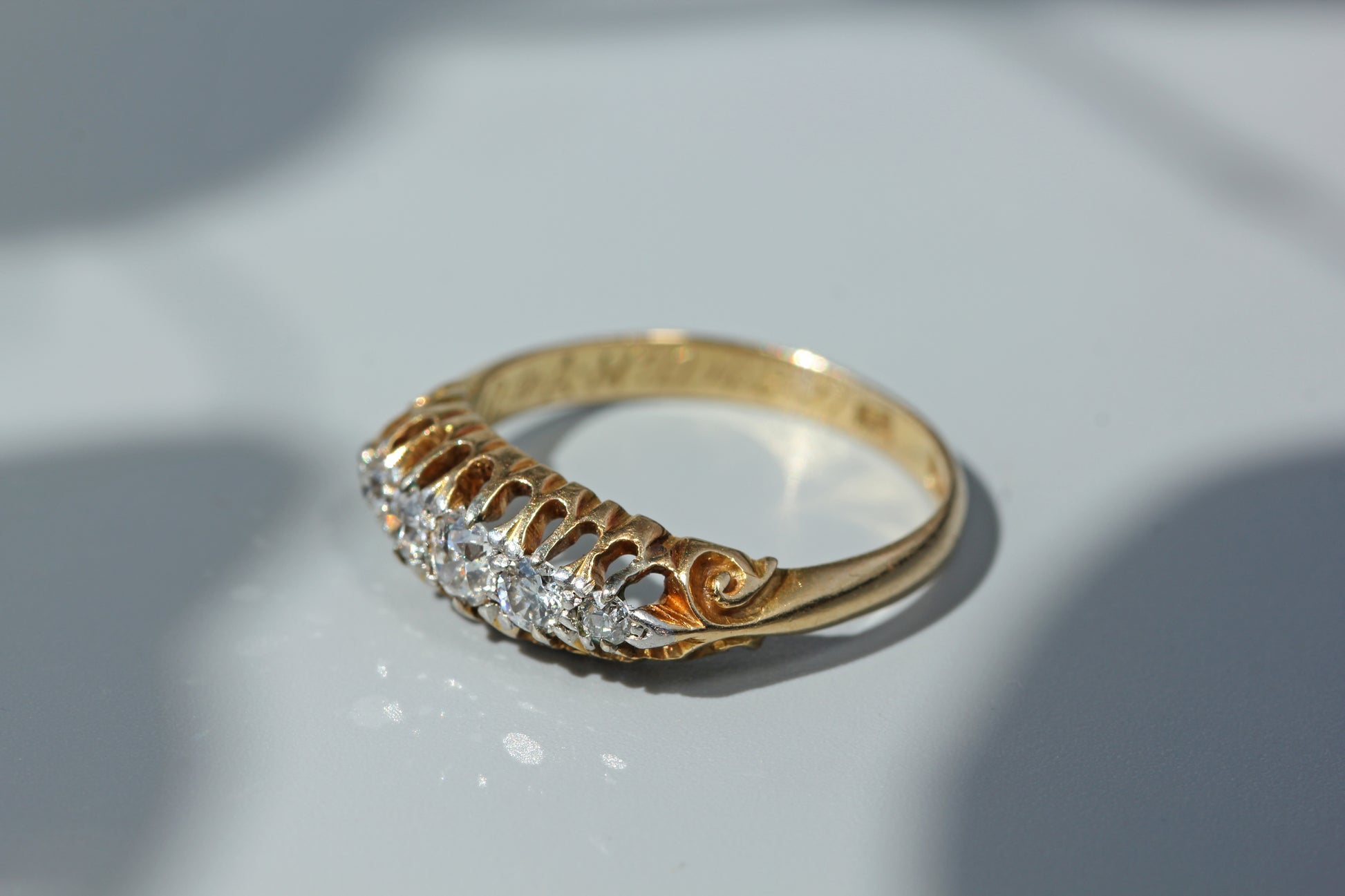 A yellow gold ring with a decorative filigree band and a row of clear gemstones on the front, photographed on a light gray surface with soft shadows.