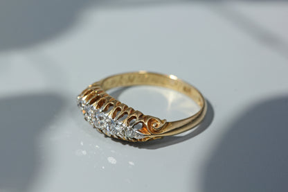 A yellow gold ring with a decorative filigree band and a row of clear gemstones on the front, photographed on a light gray surface with soft shadows.