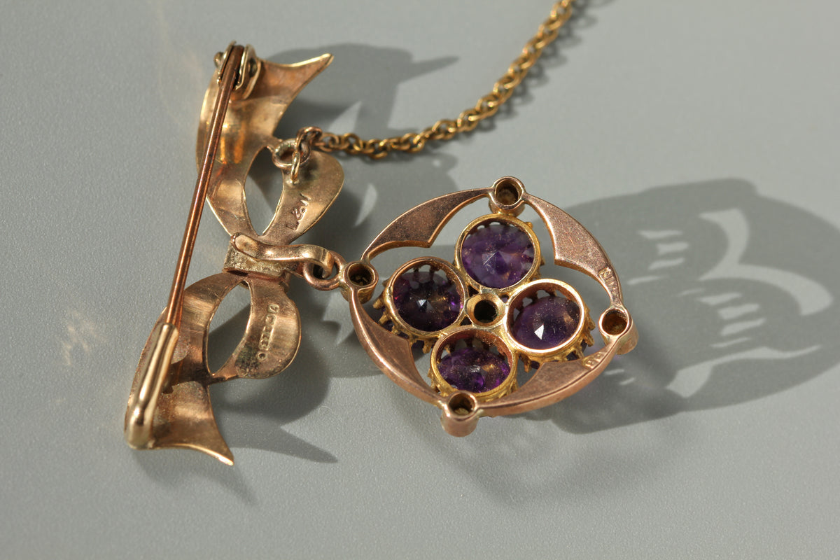 A close up of the back of a vintage 9ct gold brooch pendant, with lear hallmarks and the details of four round cut amethysts, displayed on neutral grey background 