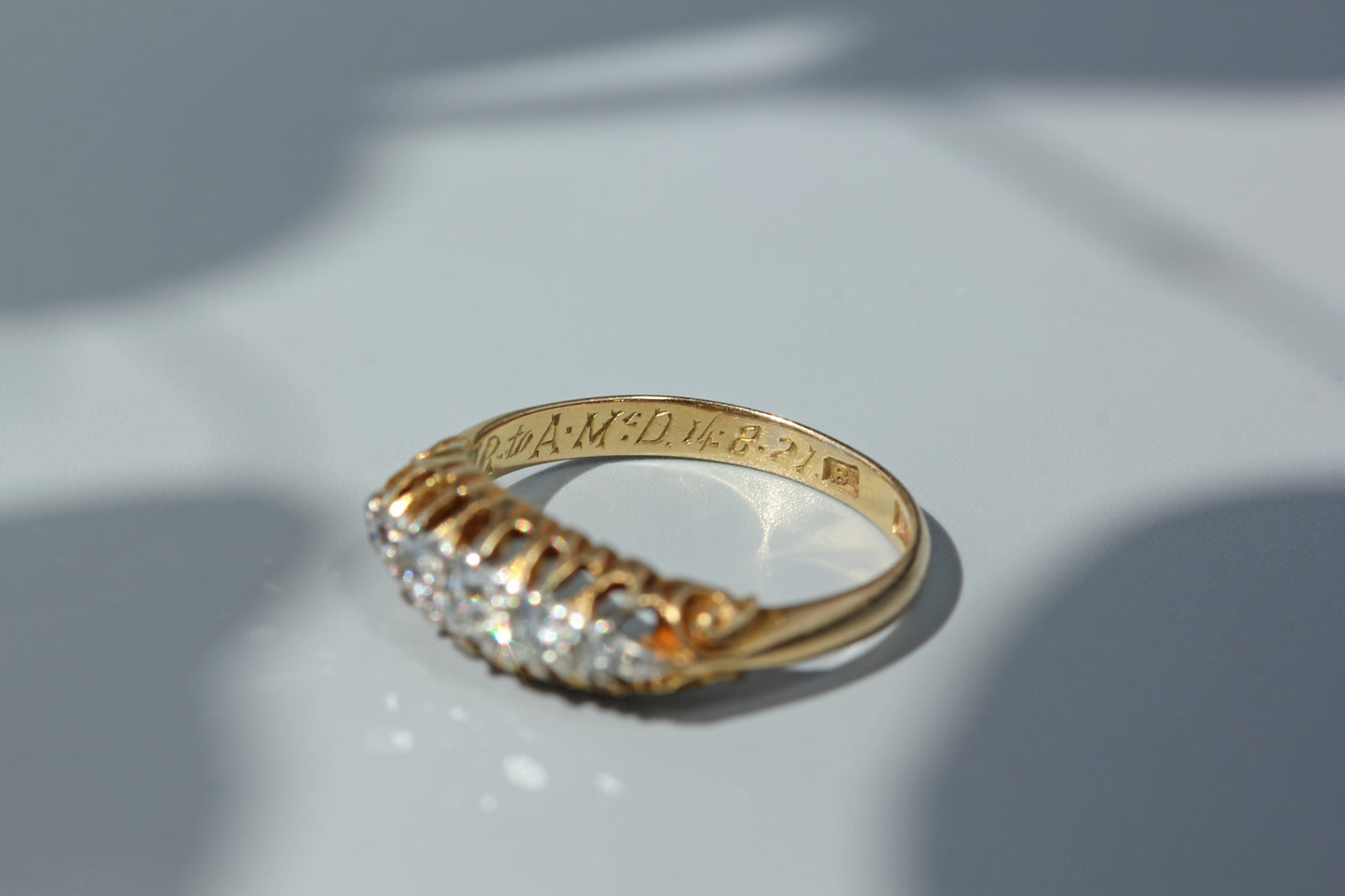 A 18ct yellow gold ring with a row of diamonds on the front, photographed on a light gray surface with soft shadows. The inside of the band is engraved with text.