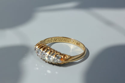 A 18ct yellow gold ring with a row of diamonds on the front, photographed on a light gray surface with soft shadows. The inside of the band is engraved with text.