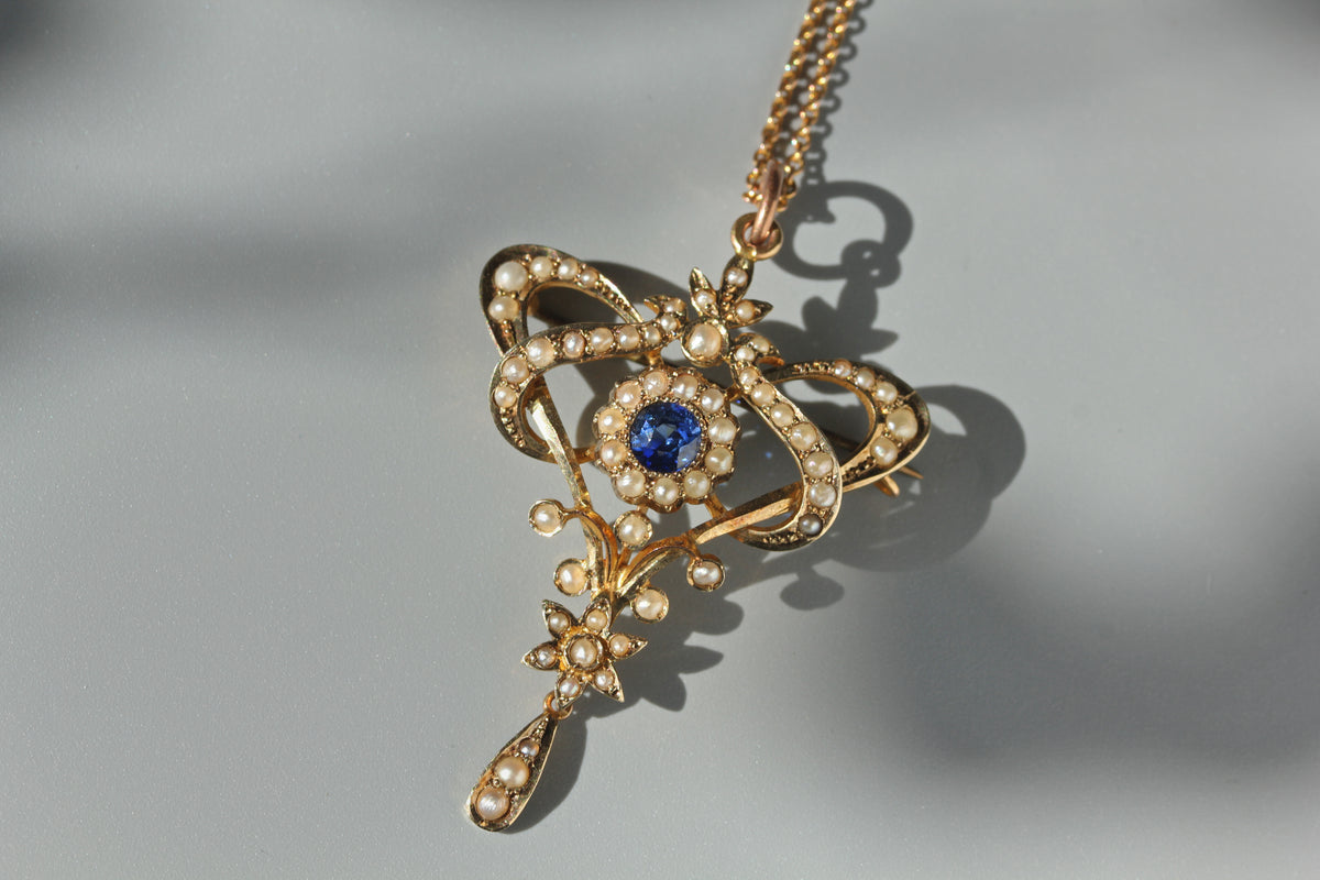 An ornate antique 9ct gold brooch pendant necklace featuring a central round blue gemstone surrounded by a halo of pearls, photographed on a soft gray background with a sharp shadow.