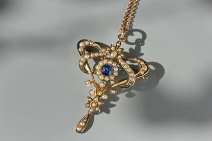 An ornate antique 9ct gold brooch pendant necklace featuring a central round blue gemstone surrounded by a halo of pearls, photographed on a soft gray background with a sharp shadow.