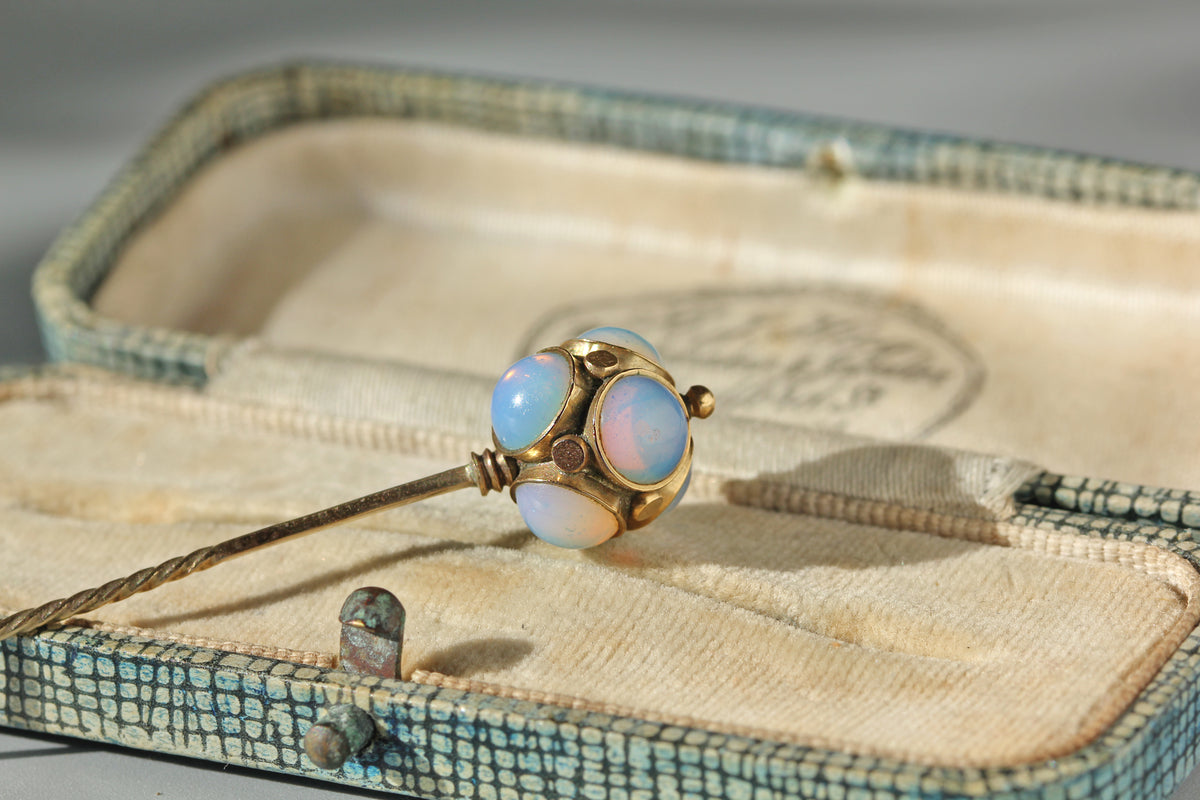An antique edwardian 9ct gold stick pin with round iridescent opal cabochons, displayed inside an open textured blue-green and beige jewelry box.