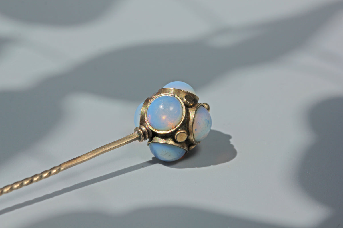 A close-up of an antique 9ct gold stick pin or lapel pin with round light blue opal like gemstones, resting on a grey surface with soft shadows.