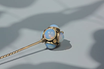A close-up of an antique 9ct gold stick pin or lapel pin with round light blue opal like gemstones, resting on a grey surface with soft shadows.