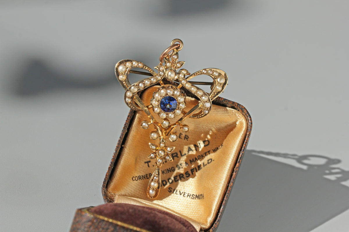 An antique Victorian pendant brooch in yellow gold and white pearls, featuring a central blue spinel, displayed inside an open vintage box with gold-lined velvet interior.