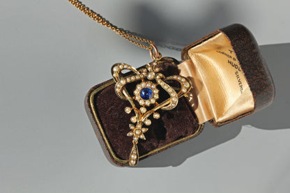 An ornate 9ct gold pendant necklace with a central blue gemstone and a halo of pearls, displayed on an open vintage presentation box against a neutral grey background.