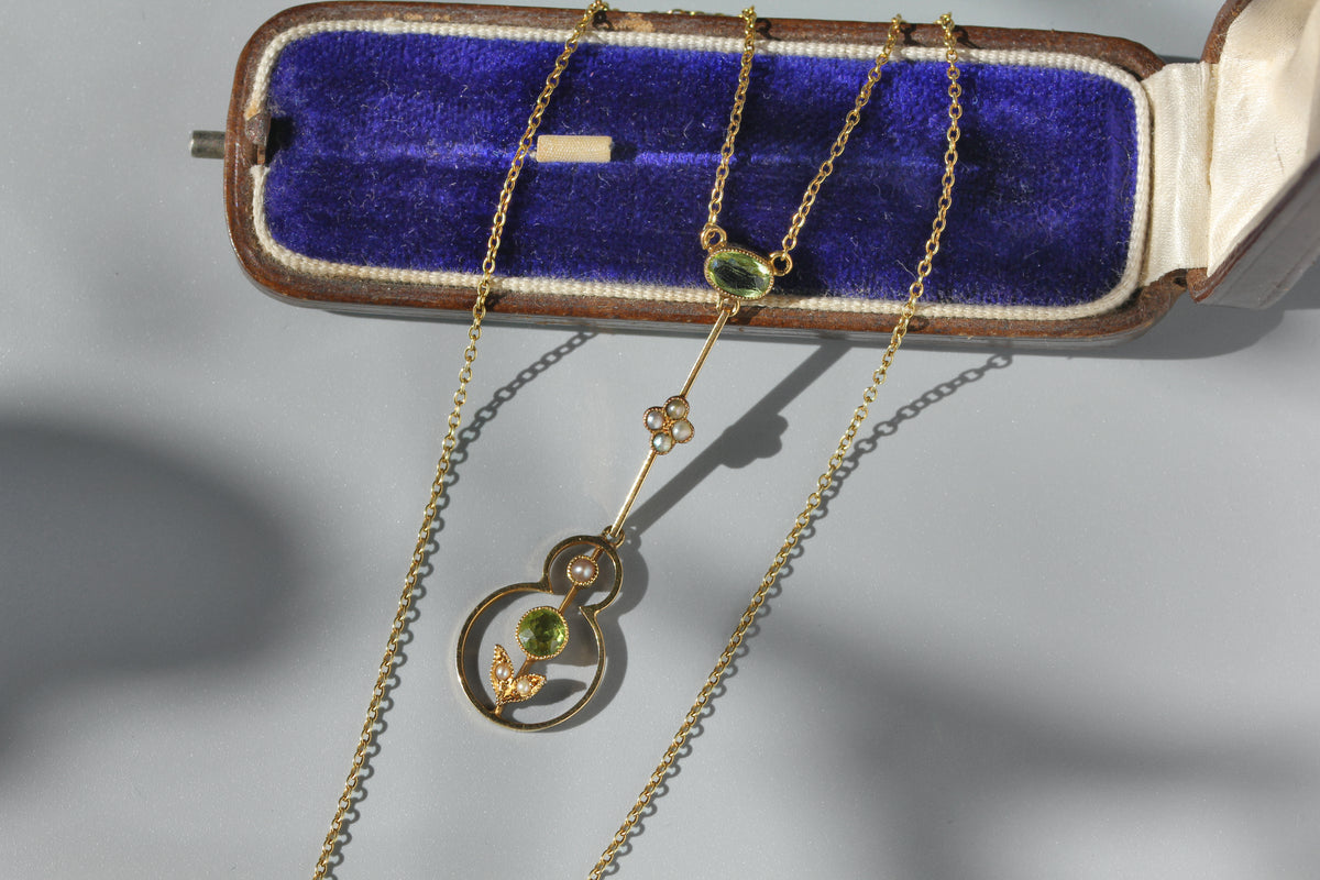 Victorian antique 15ct gold necklace with a circular pendant featuring a green peridot stone and pearl flowers, displayed on an open blue velvet jewelry box against a light gray background