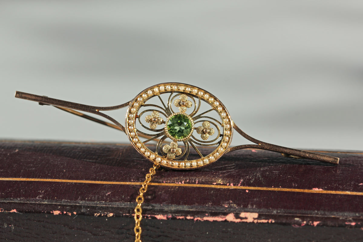 An antique edwardian 9ct gold brooch pin with a circular filigree center, a green peridot gemstone and a halo of small seed pearls, resting on a dark wooden jewellery box.