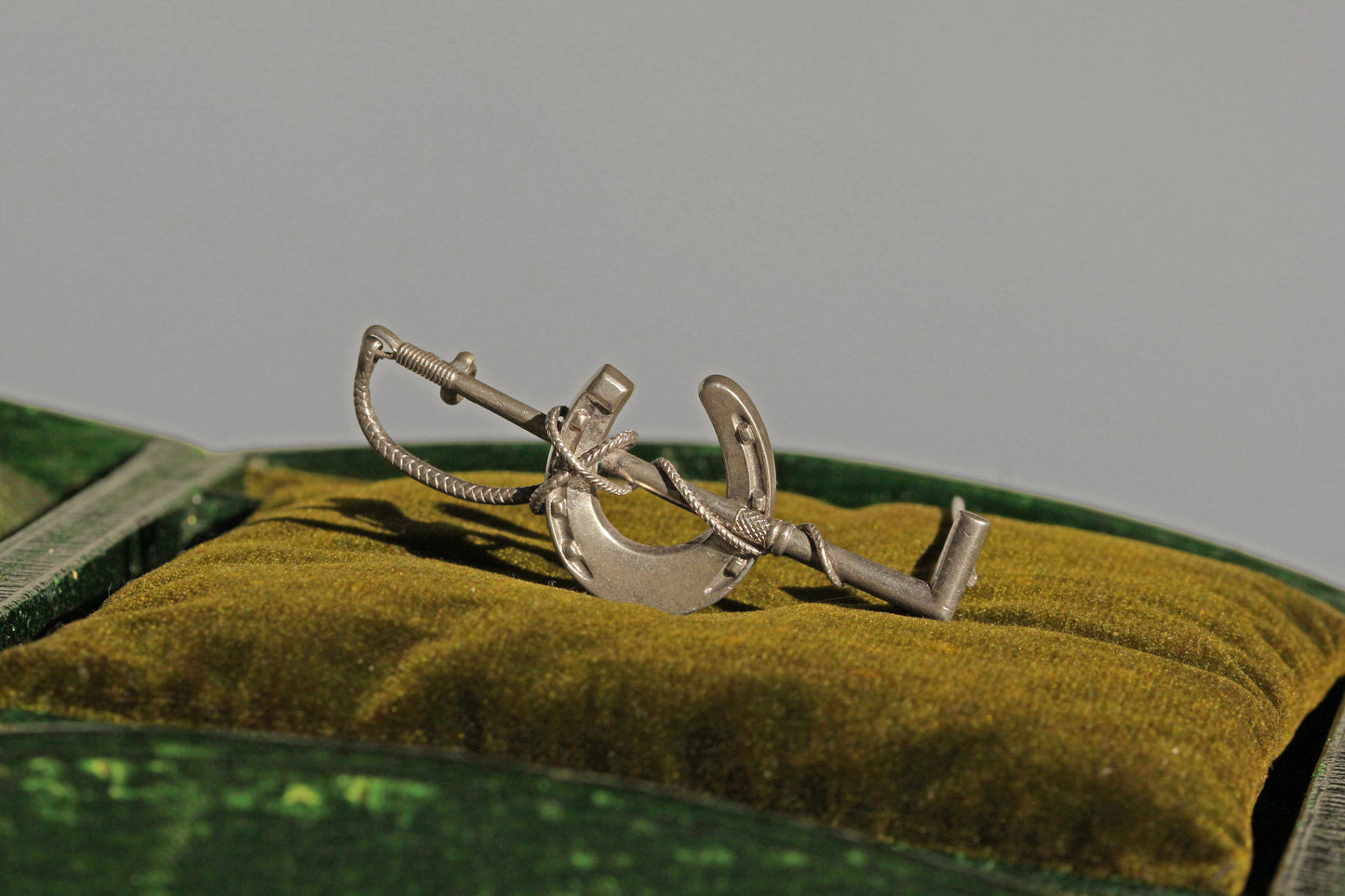 Antique sterling silver horseshoe and riding crop brooch displayed on olive velvet cushion
