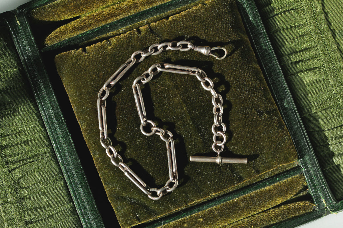 Antique sterling silver fancy link watch chain with dog clip and t bar, displayed on a green velvet antique jewellery box 