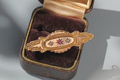 An ornate victorian 9ct brooch with star motifs and a central pink gemstone with two diamonds either side, displayed inside an open vintage jewelry box with a dark brown velvet interior.