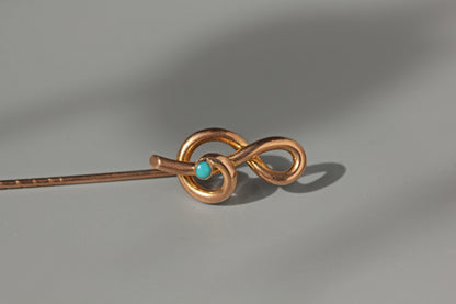 A copper-colored metal hair pin with a twisted infinity loop design and a small round turquoise stone, shown on a neutral gray background.