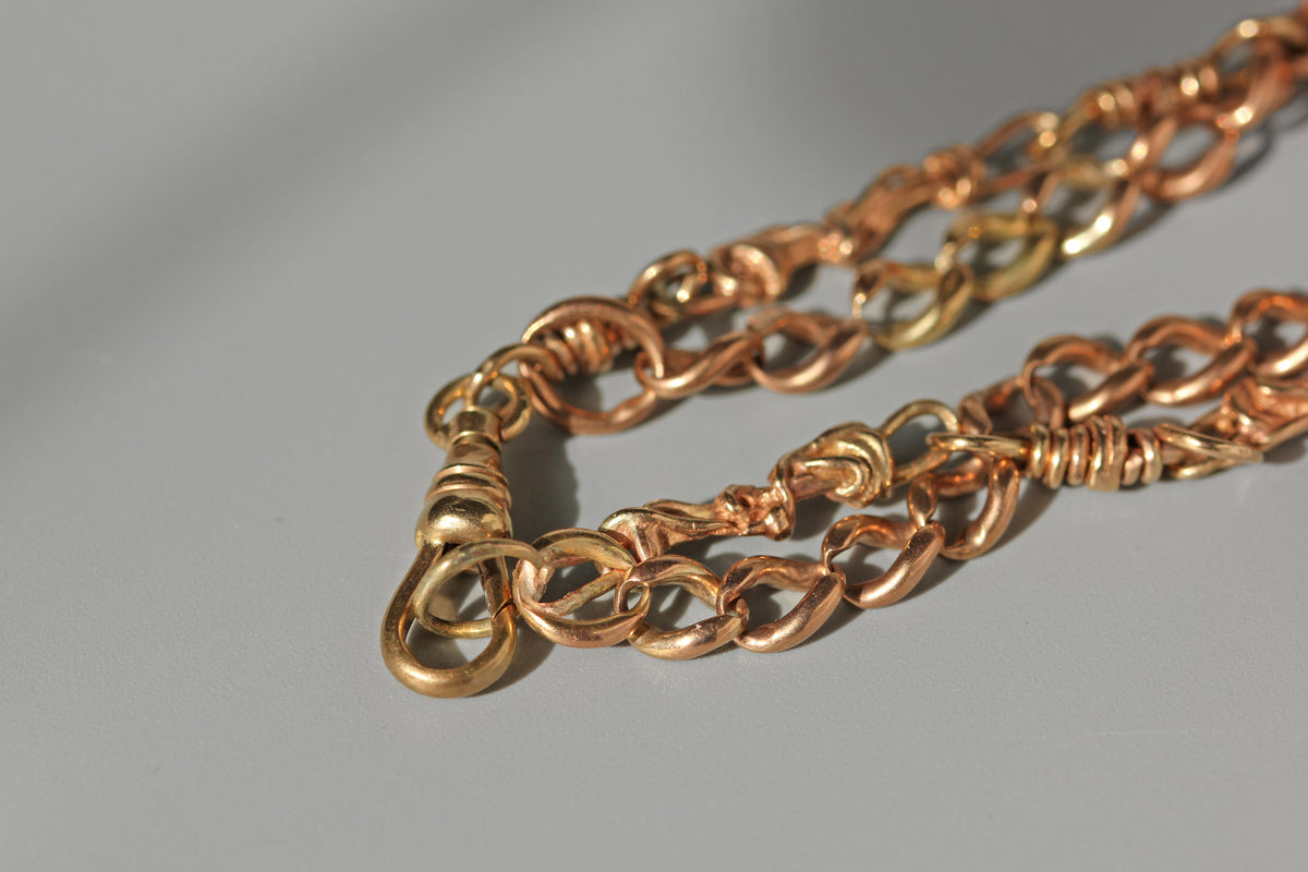 A close-up of a chunky chain necklace with alternating polished gold and rose gold links, resting on a neutral gray surface.