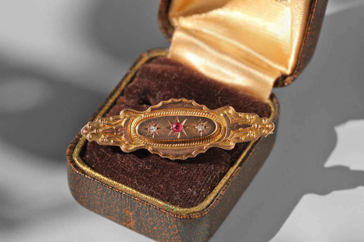 An ornate vintage gold oval brooch with a central red gemstone and star motifs, displayed inside an open brown velvet jewelry box with a gold satin lining.