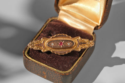 An ornate vintage gold oval brooch with a central red gemstone and star motifs, displayed inside an open brown velvet jewelry box with a gold satin lining.