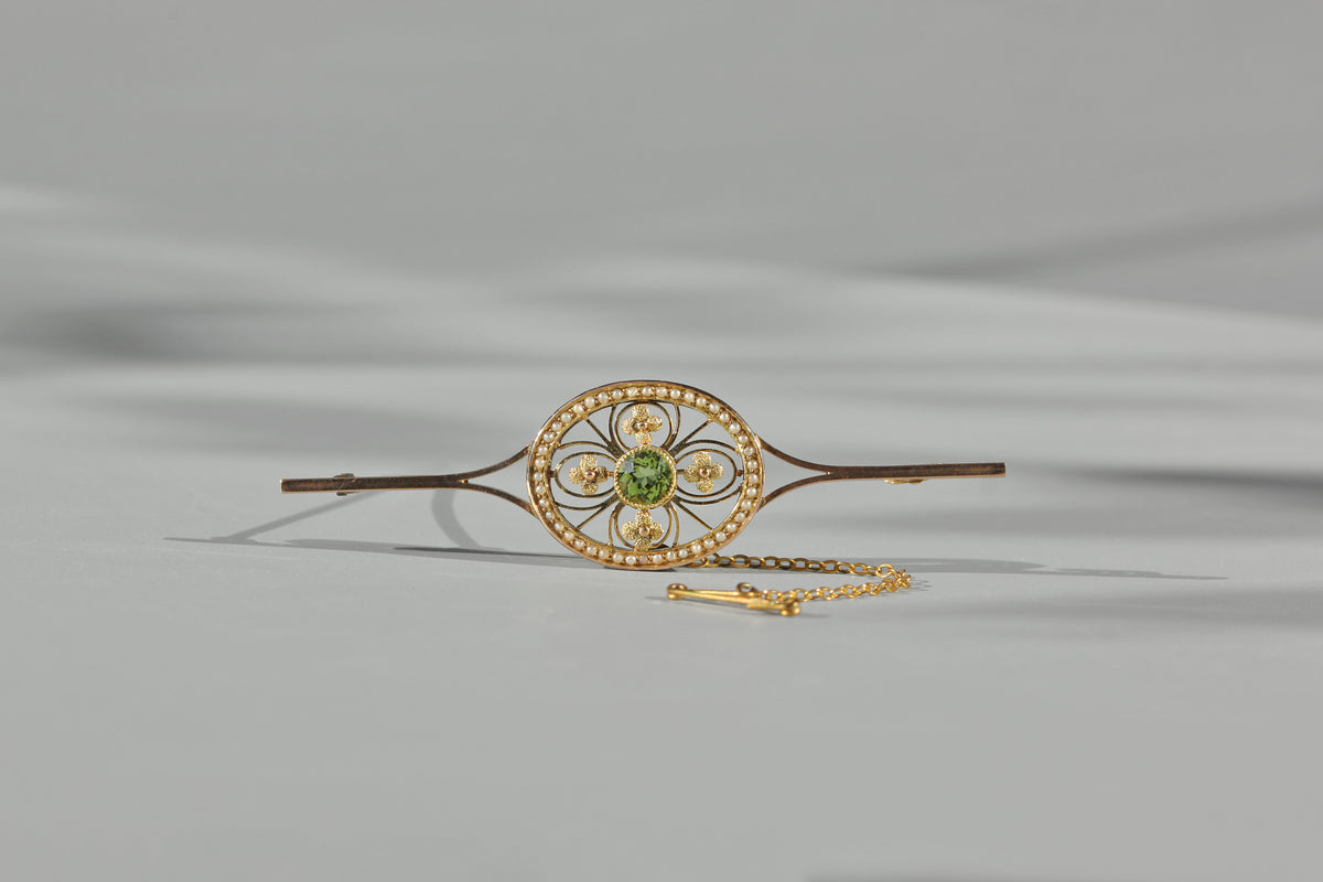 An antique edwardian brooch made of 9ct yellow gold, featuring a central green peridot gemstone surrounded by smaller gold and pearl accents, with a delicate gold chain attached, set against a neutral gray background.