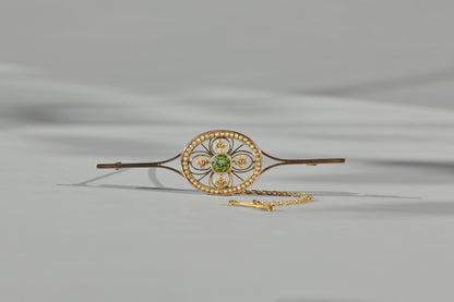 An antique edwardian brooch made of 9ct yellow gold, featuring a central green peridot gemstone surrounded by smaller gold and pearl accents, with a delicate gold chain attached, set against a neutral gray background.