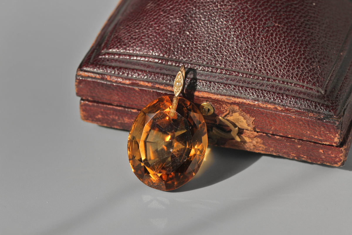 An antique oval faceted amber-yellow citrine gemstone pendant with a gold bail, resting in front of an antique dark brown leather case on a light gray surface.