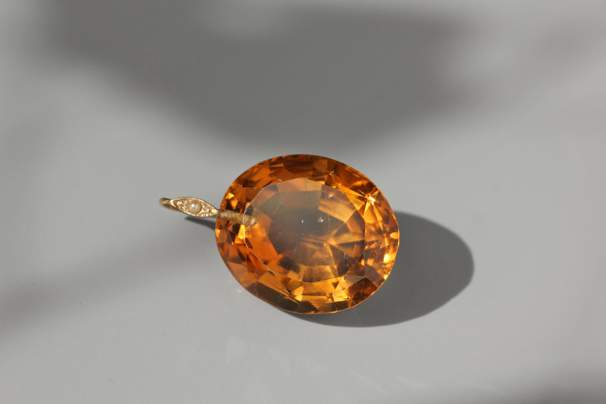 An oval-cut orange gemstone pendant set in a gold-toned bezel with a small diamond accent, photographed on a light gray surface with a soft shadow.