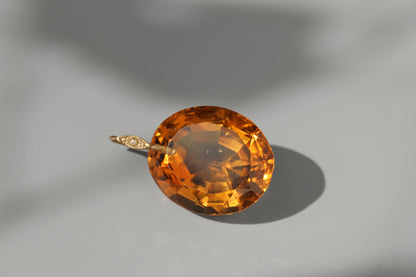 An oval-cut orange gemstone pendant set in a gold-toned bezel with a small diamond accent, photographed on a light gray surface with a soft shadow.