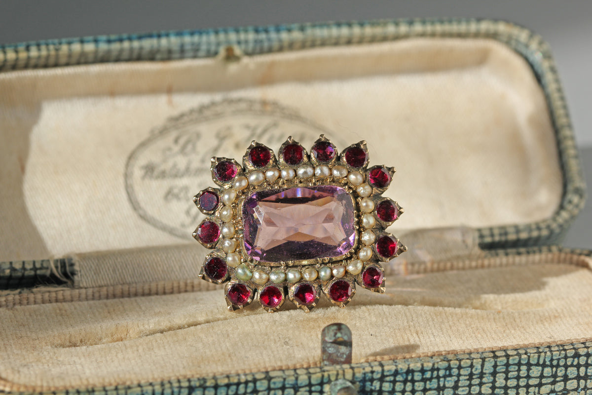 A 9ct gold antique brooch from the Georgian era with a cushion cut amethyst surrounded by seed pearls and red flat cut garnets in a closed back setting, displayed inside an antique blue-green jewelry box with cream lining
